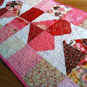 May include: A pink and red patchwork quilt table runner with a quilted design. The runner features a variety of floral and geometric patterns.