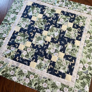 Op de afbeelding: Een quilt met een patchwork-ontwerp met een groene achtergrond met witte bloemen en vlinders. De quilt is gemaakt van vierkanten in verschillende kleuren, waaronder geel, blauw en wit. De quilt is afgewerkt met een witte rand.