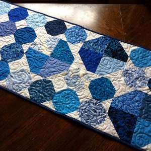May include: A blue and white quilted table runner with a geometric pattern. The runner is made of fabric with a swirling design and has a dark blue border.