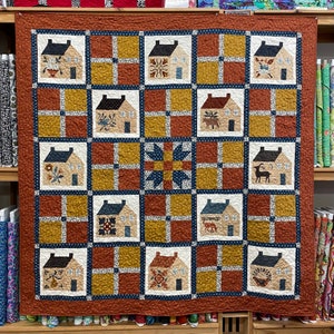 May include: A quilt with a patchwork design featuring a grid of 16 squares. Each square contains a different image of a house with various details, including flowers, birds, and stars. The quilt is made with a variety of fabrics in shades of brown, blue, and yellow.