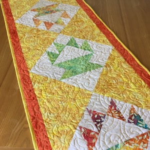 May include: A handmade, quilted table runner with a vibrant design. The runner features a yellow background with a floral pattern, accented by orange and white geometric shapes. The edges are trimmed with orange fabric.