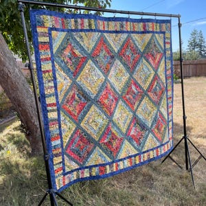 May include: A square quilt featuring a diamond-patterned patchwork design in shades of red, yellow, and blue. The quilt is bordered by a dark blue fabric frame and is suspended from a black metal stand, showcasing intricate fabric textures.