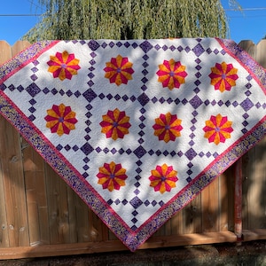 Può includere: Una trapunta fatta a mano con sfondo bianco con un motivo ripetuto di forme floreali arancioni e rosa e accenti quadrati viola. La trapunta ha un bordo decorativo con un motivo floreale ed è esposta all'aperto.