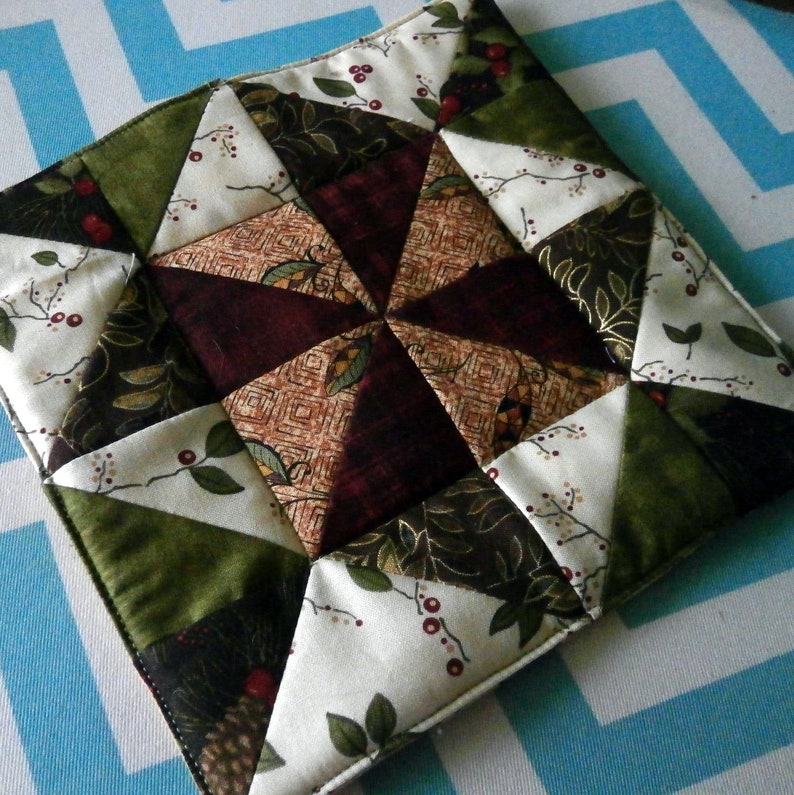 May include: A quilted fabric pot holder with a starburst design in shades of green, brown, and white. The fabric features a floral pattern with berries and leaves.