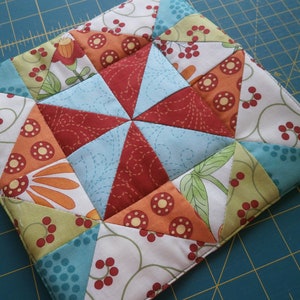 May include: A colorful patchwork quilt square with a starburst design in the center. The square is made of various fabrics with floral and geometric patterns in shades of red, orange, green, blue, and white.