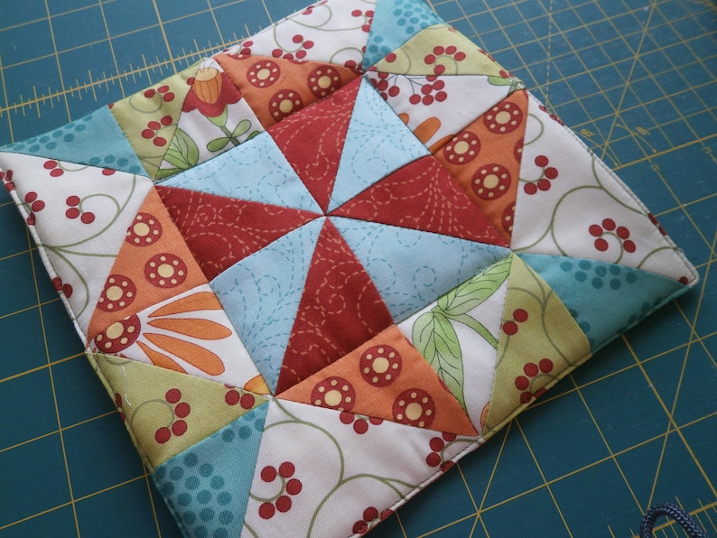 May include: A colorful patchwork quilt square with a starburst design in the center. The square is made of various fabrics with floral and geometric patterns in shades of red, orange, green, blue, and white.