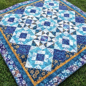 May include: A square quilt with a geometric pattern in shades of blue, white, and purple. The quilt features a central design of squares and triangles, bordered by floral and gold accents. The quilt is displayed on a green grassy surface.