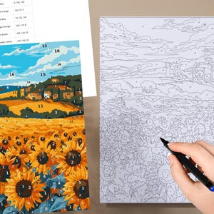 May include: A paint-by-numbers kit depicting a sunflower field and a Tuscan landscape. The kit includes a canvas with numbered sections, paint colours, and a reference sheet. A hand is shown holding a black pen, ready to paint.