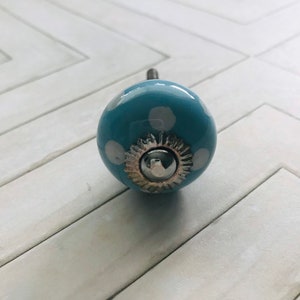 May include: A round, light blue ceramic cabinet knob featuring white polka dots. The center of the knob is adorned with a silver-toned metal decorative plate and a small, smooth, circular silver-toned metal cap at the very front.