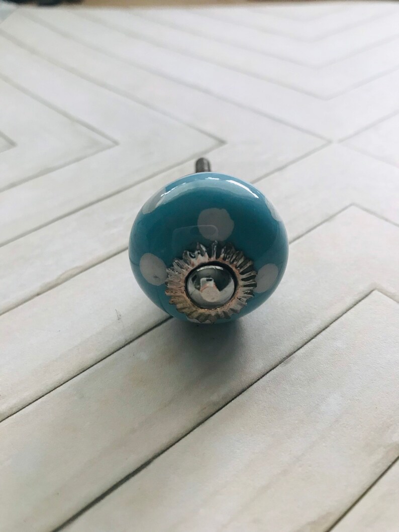 May include: A round, light blue ceramic cabinet knob featuring white polka dots. The center of the knob is adorned with a silver-toned metal decorative plate and a small, smooth, circular silver-toned metal cap at the very front.