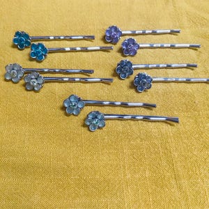 May include: A collection of flower-shaped bobby pins in various colors, including blue, purple, and silver. The bobby pins have a silver metal base and are arranged on a yellow fabric surface. These hair accessories are ideal for adding a touch of floral elegance.