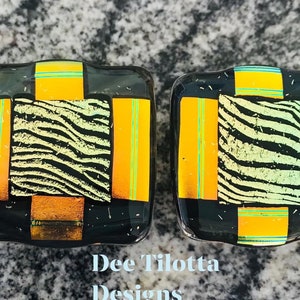 May include: Two square glass decorative pieces with a black base, featuring orange, yellow, and green accents. One section has a zebra stripe pattern. The text "Dee Tilotta Designs" is visible.