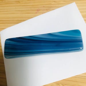 May include: A rectangular hair clip featuring a smooth, glossy finish with horizontal, swirling bands of various shades of blue, ranging from deep navy to light cyan, creating a fluid, layered appearance across the entire surface.