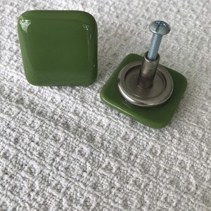 May include: Two olive green square cabinet knobs. One knob is complete, with a silver screw and backing plate. The other knob is shown separately. The knobs have a glossy finish and are likely made of glass or resin.