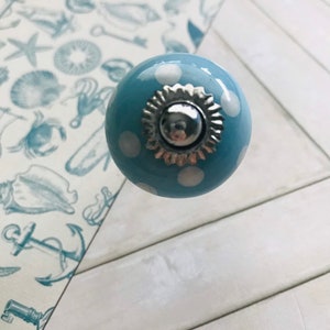 May include: A round, light blue ceramic cabinet knob featuring white polka dots. The center of the knob is adorned with a decorative, scalloped silver-toned metal cap that has a polished, reflective finish at its very peak.