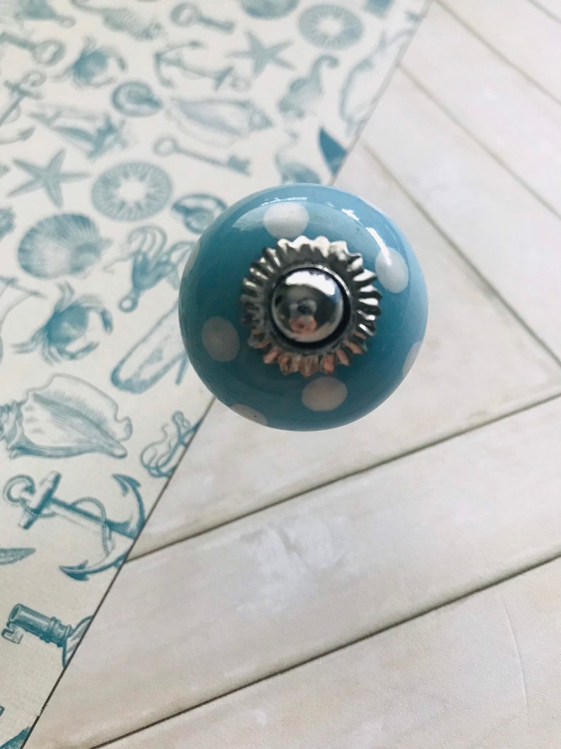 May include: A round, light blue ceramic cabinet knob featuring white polka dots. The center of the knob is adorned with a decorative, scalloped silver-toned metal cap that has a polished, reflective finish at its very peak.