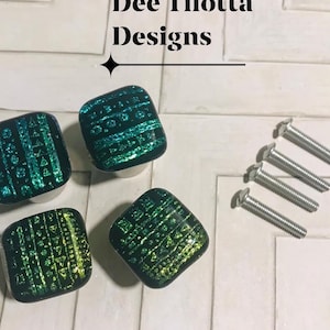 May include: Four square cabinet knobs with a black base and iridescent green and blue geometric patterns. The text Dee Tilotta Designs is visible above the knobs. Four silver metal mounting screws are included with the set.