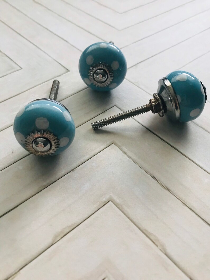 May include: Three round cabinet knobs with a light blue finish and white polka dot pattern. Each knob features a central silver-colored metal cap with a sunburst-style edge and a threaded metal bolt extending from the back for installation.