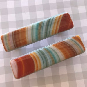 May include: Two rectangular hair barrettes with a striped pattern in shades of brown, orange, teal, and white. The barrettes have rounded edges and a glossy finish. They are displayed on a gray and white checkered surface.