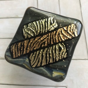 May include: A dichroic glass art piece in a diamond shape. The glass is a dark gray with gold and bronze zebra stripe designs. The edges are rounded and the glass has a glossy finish.