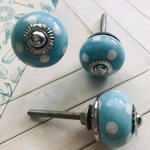May include: Three round ceramic cabinet knobs featuring a light blue base with white polka dots. Each knob has a silver-toned metal base and a central decorative silver-toned metal cap with a scalloped edge. A threaded metal bolt extends from the back.