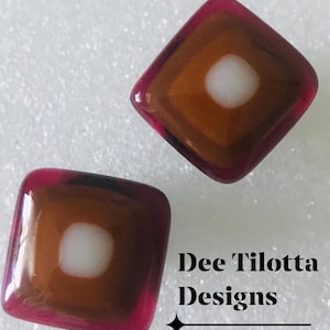 May include: Two square-shaped glass beads. Each bead has a dark red border, a brown center, and a white dot in the middle. The text "Dee Tilotta Designs" is visible.