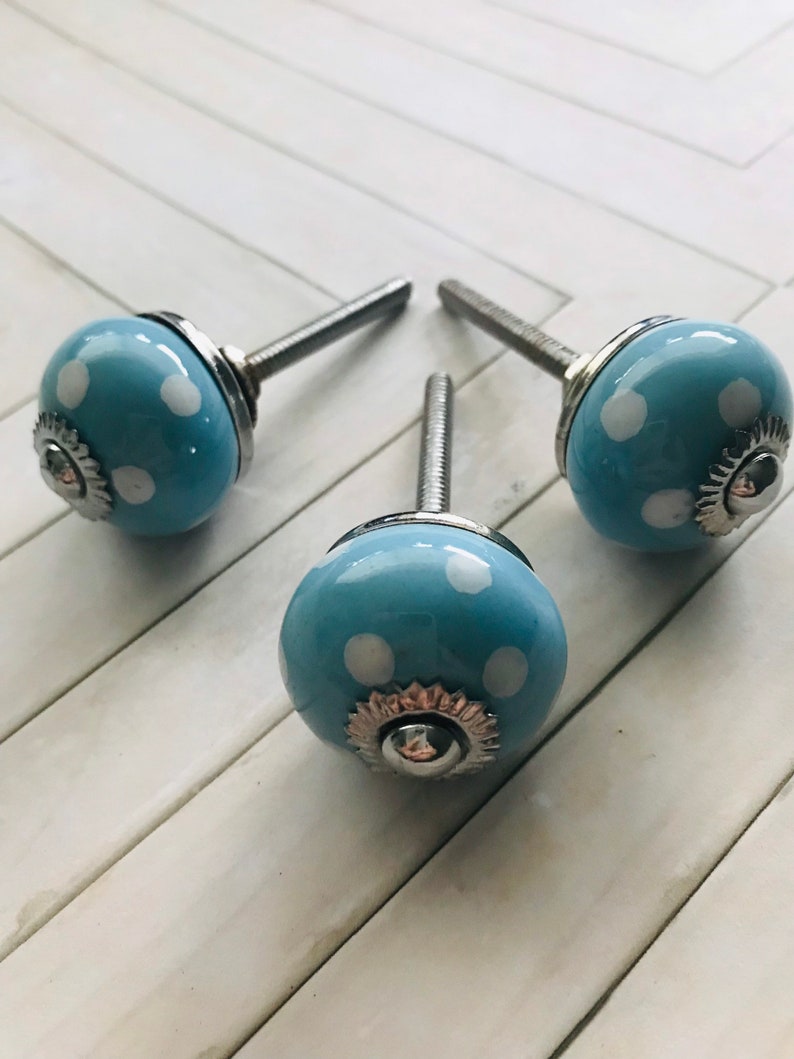 May include: Three round ceramic cabinet knobs in a light blue color with white polka dots. Each knob features a silver-toned metal base and a threaded bolt extending from the back, with a small silver-toned decorative cap at the center front.