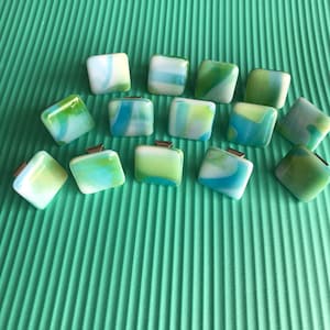 May include: A collection of square glass pendants with a marbled effect in shades of blue, green, and white. Each pendant has a small silver-colored bail attached. The pendants are arranged on a textured, light green surface.