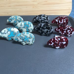 May include: A collection of decorative glass animal figurines in various colors. The figurines are shaped like small animals, with patterns in shades of blue, white, black, gray, and red. They are arranged on a gray surface.