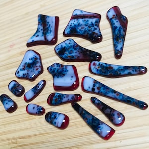 May include: A collection of colorful glass pieces in various shapes. The glass features a combination of red, white, and blue speckled patterns. The pieces are arranged on a light brown wooden surface.