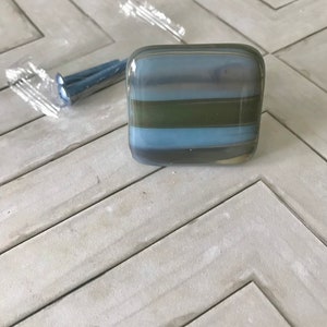 May include: A square glass cabinet knob with horizontal stripes of blue, green, and gray. The knob is next to a package of screws. The knob is likely for a drawer or cabinet.