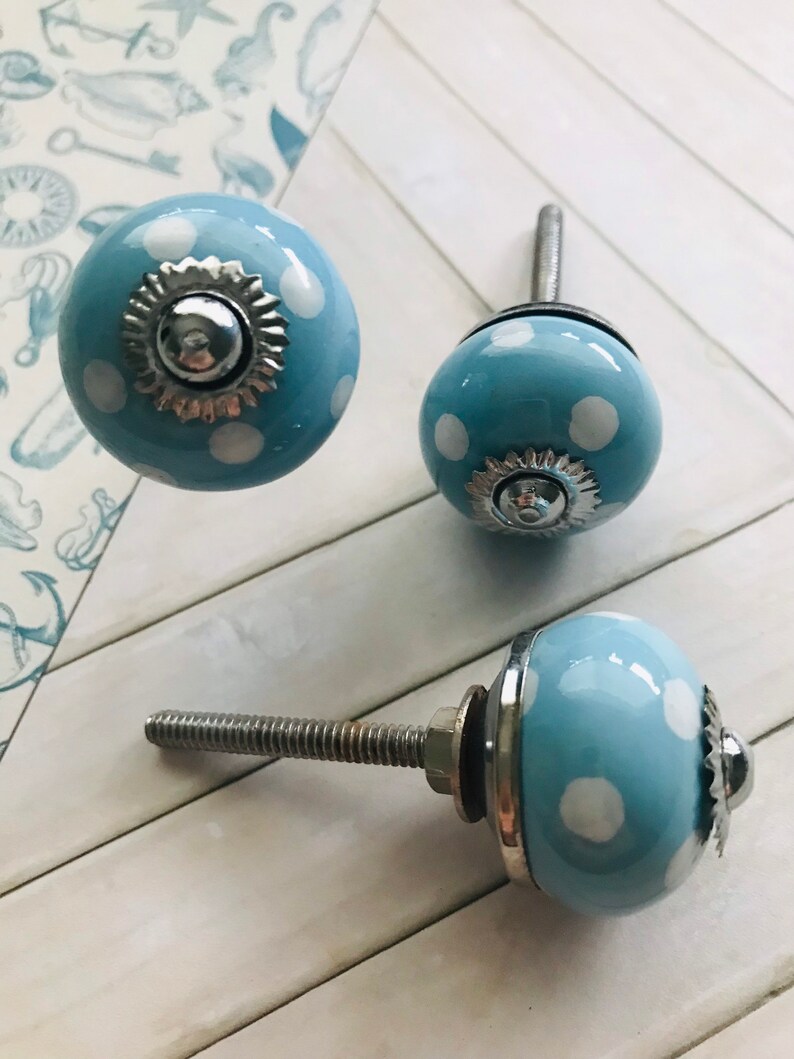 May include: Three round ceramic cabinet knobs featuring a light blue base with white polka dots. Each knob has a silver-toned metal base and a central decorative silver-toned metal cap with a scalloped edge. A threaded metal bolt extends from the back.
