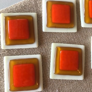 May include: Close-up of several square glass tiles. Each tile has a white border, a golden-yellow middle layer, and a bright orange square center. The tiles are arranged on a textured, neutral-colored surface.