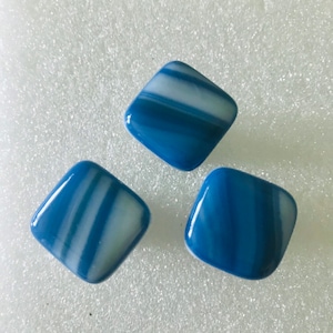 May include: Three square, blue glass decorative tiles with white and blue striped patterns. The tiles have rounded corners and a glossy finish. The tiles are arranged on a white surface.