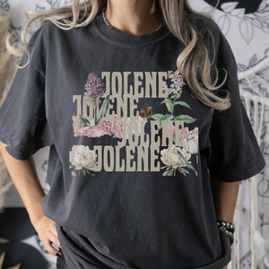 Comfort colors shirt in the color Pepper with the word Jolene in a staggered and stacked beige font written 4 times. There are wildflowers surrounding the words. Pepper is a Dark Gray