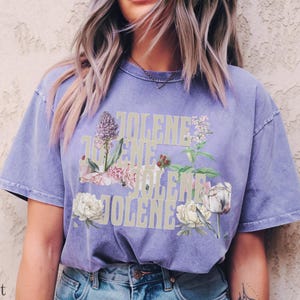 Op de afbeelding: Een violetkleurig T-shirt met korte mouwen waarop het woord JOLENE in een groot, lichtgekleurd bloklettertype is gedrukt. De tekst is bedekt met gedetailleerde botanische illustraties van diverse bloemen, waaronder witte pioenrozen en paarse bloemstengels.