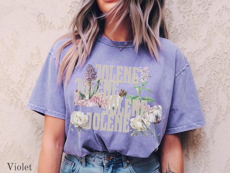 Op de afbeelding: Een violetkleurig T-shirt met korte mouwen waarop het woord JOLENE in een groot, lichtgekleurd bloklettertype is gedrukt. De tekst is bedekt met gedetailleerde botanische illustraties van diverse bloemen, waaronder witte pioenrozen en paarse bloemstengels.