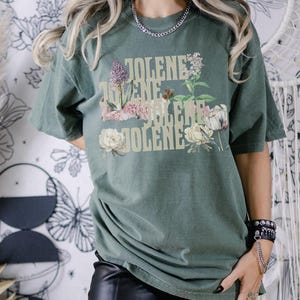 Comfort colors shirt in the color Moss with the word Jolene in a staggered and stacked beige font written 4 times. There are wildflowers surrounding the words. Moss is a muted green color