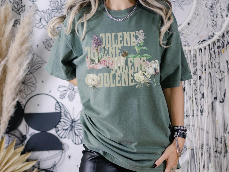 Comfort colors shirt in the color Moss with the word Jolene in a staggered and stacked beige font written 4 times. There are wildflowers surrounding the words. Moss is a muted green color