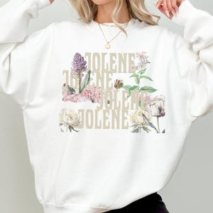 Op de afbeelding: Een wit sweatshirt met ronde hals en het woord JOLENE in een groot, lichtbeige schreefloos lettertype. De tekst is bedekt met een kleurrijke botanische illustratie van diverse bloemen, waaronder roze, witte en paarse bloesems.