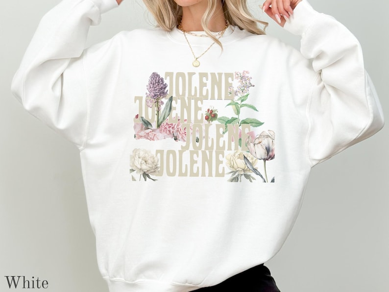 Op de afbeelding: Een wit sweatshirt met ronde hals en het woord JOLENE in een groot, lichtbeige schreefloos lettertype. De tekst is bedekt met een kleurrijke botanische illustratie van diverse bloemen, waaronder roze, witte en paarse bloesems.