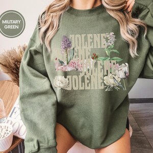 Op de afbeelding: Een militair groene sweater met ronde hals en de herhaalde tekst JOLENE in een lichtbeige lettertype, gelaagd met botanische illustraties van paarse, roze en witte bloemen en groen blad. Een ronde badge in de hoek vermeldt MILITARY GREEN.