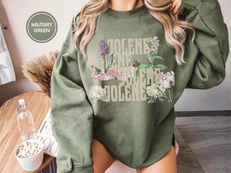 Op de afbeelding: Een militair groene sweater met ronde hals en de herhaalde tekst JOLENE in een lichtbeige lettertype, gelaagd met botanische illustraties van paarse, roze en witte bloemen en groen blad. Een ronde badge in de hoek vermeldt MILITARY GREEN.