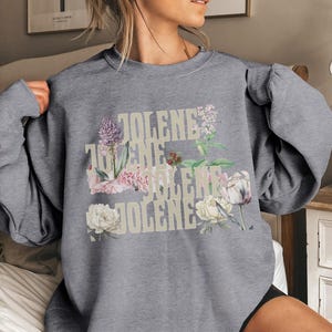 Op de afbeelding: Een grijs gem&ecirc;leerd sweatshirt met ronde hals en de herhaalde tekst JOLENE in een lichtbeige lettertype. Het ontwerp is gelaagd met botanische illustraties van diverse bloemen, waaronder witte pioenrozen, paarse alliums en groen blad.