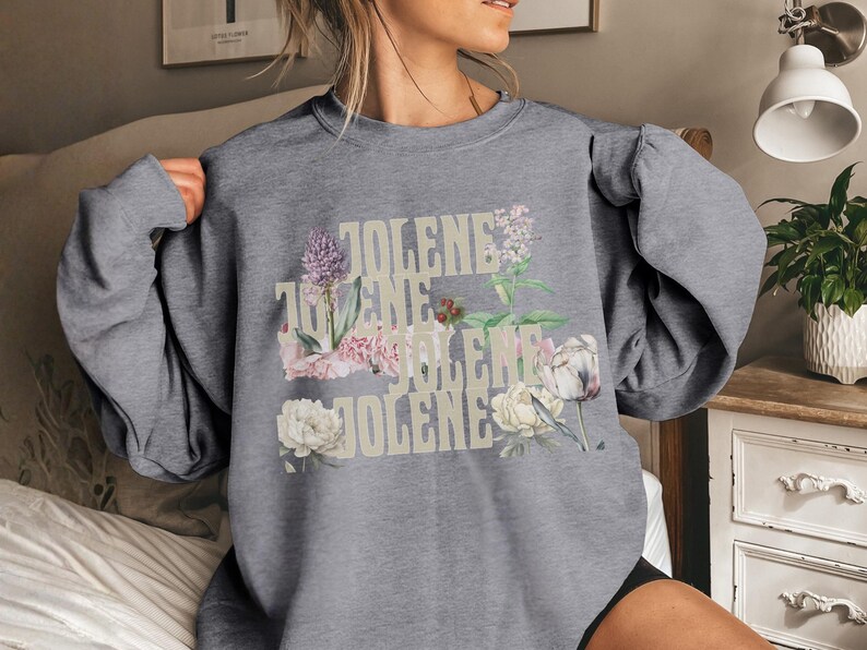 Op de afbeelding: Een grijs gem&ecirc;leerd sweatshirt met ronde hals en de herhaalde tekst JOLENE in een lichtbeige lettertype. Het ontwerp is gelaagd met botanische illustraties van diverse bloemen, waaronder witte pioenrozen, paarse alliums en groen blad.