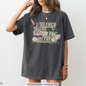 Op de afbeelding: Een antracietgrijs T-shirt met korte mouwen en de tekst JOLENE in een gestapelde lay-out, geaccentueerd met illustraties van diverse bloemen, waaronder pioenrozen en paarse bloemen. Het T-shirt heeft een ronde hals en een relaxte pasvorm.