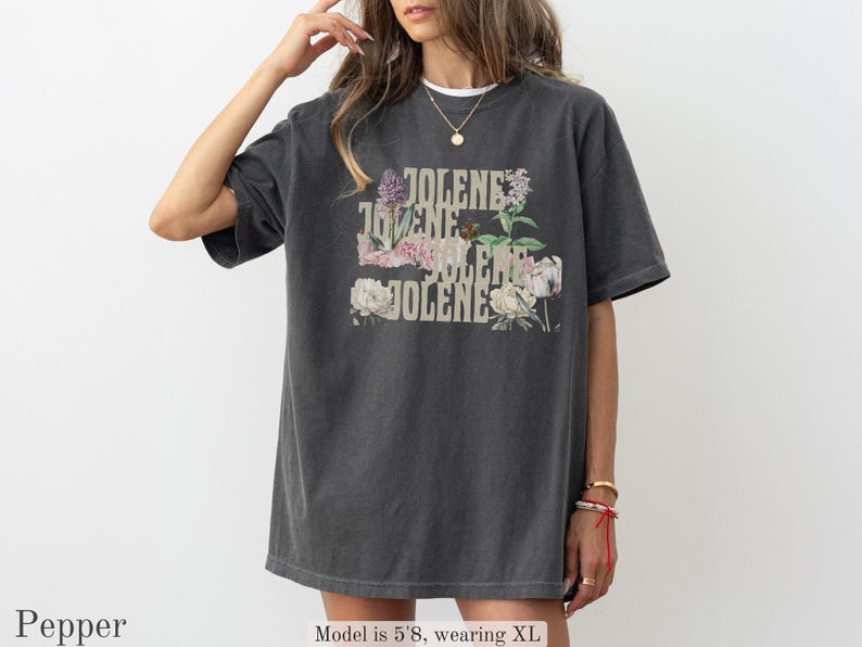 Op de afbeelding: Een antracietgrijs T-shirt met korte mouwen en de tekst JOLENE in een gestapelde lay-out, geaccentueerd met illustraties van diverse bloemen, waaronder pioenrozen en paarse bloemen. Het T-shirt heeft een ronde hals en een relaxte pasvorm.