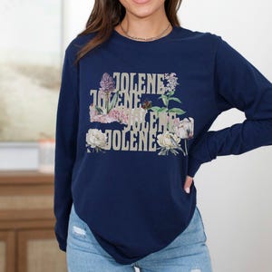 Op de afbeelding: Een marineblauw sweatshirt met lange mouwen en de tekst JOLENE vier keer herhaald in een gestapeld, verweerd lettertype. Het ontwerp is versierd met kleurrijke bloemenillustraties, waaronder paarse seringen, roze pioenrozen en witte bloemen.