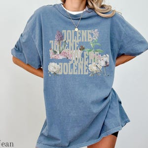 Op de afbeelding: Een jeansblauw t-shirt met korte mouwen waarop de naam JOLENE vier keer in een lichtgekleurd, gestapeld bloklettertype is gedrukt. De tekst is gelaagd met botanische illustraties van paarse en witte bloemen en groene bladeren.