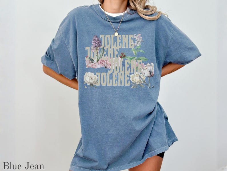 Op de afbeelding: Een jeansblauw t-shirt met korte mouwen waarop de naam JOLENE vier keer in een lichtgekleurd, gestapeld bloklettertype is gedrukt. De tekst is gelaagd met botanische illustraties van paarse en witte bloemen en groene bladeren.